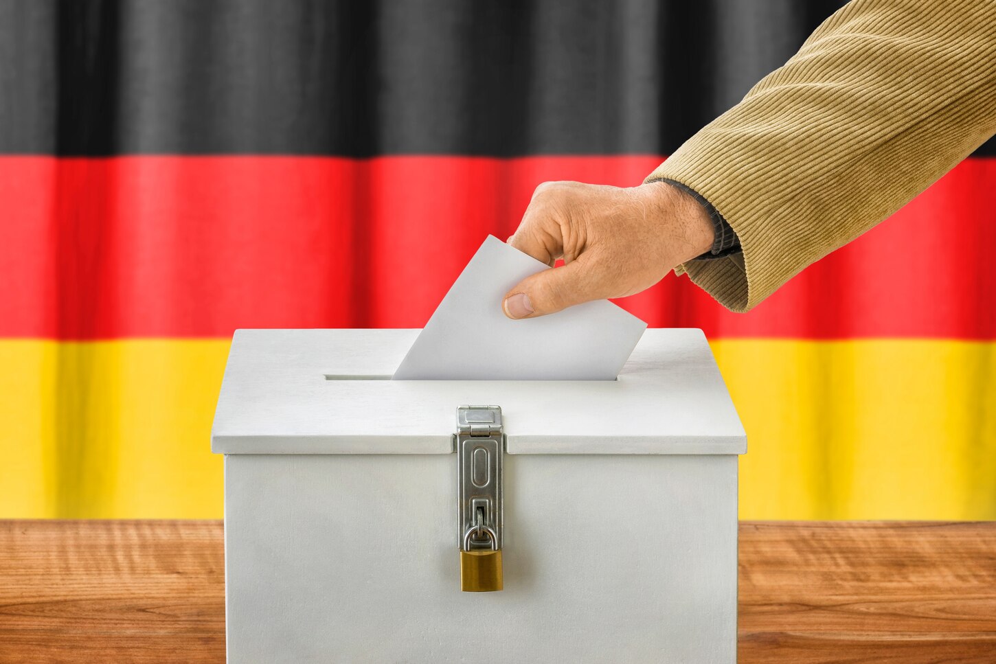 Das Bild zeigt eine Hand die einen Stimmzettel in eine weiße Wahlurne wirft, die vor einer Deutschlandfahne steht.